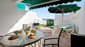 Un tavolo rotondo in vetro con cibi per la colazione, posti a sedere all'aperto con sedie e un ombrellone verde, e vista sul mare.
