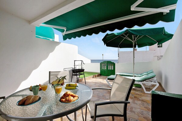 A round glass table with breakfast items, outdoor seating with chairs and a green umbrella, and a view of the sea.