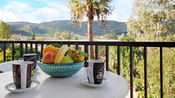 Une table avec un panier de fruits et deux tasses à café, placée sur un balcon donnant sur un paysage montagneux.