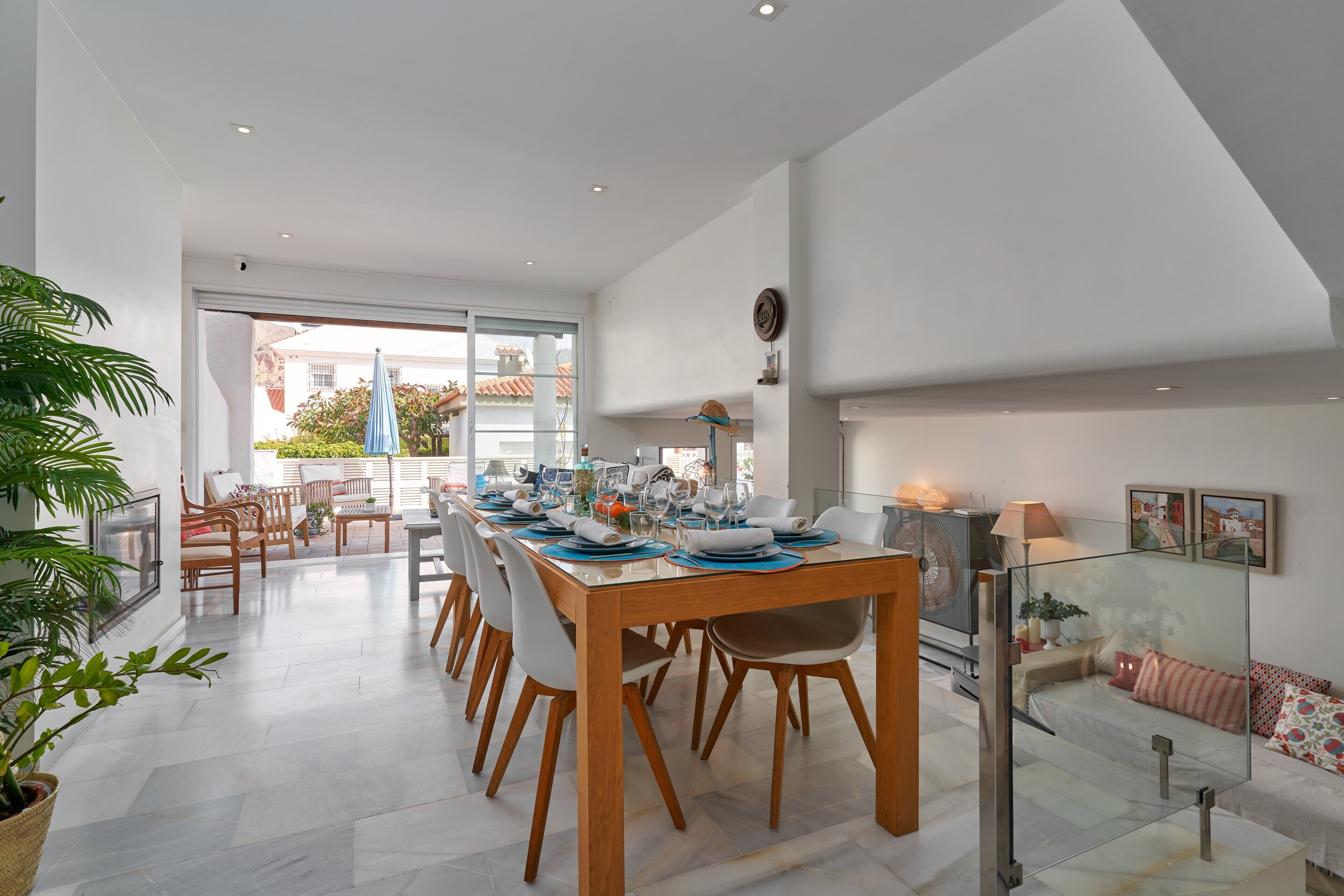 A modern dining area with a wooden table set for four, surrounded by chairs, with a glass partition and a sofa with cushions in the background.
