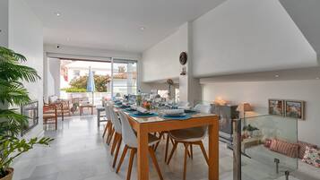 A modern dining area with a wooden table set for four, surrounded by chairs, with a glass partition and a sofa with cushions in the background.