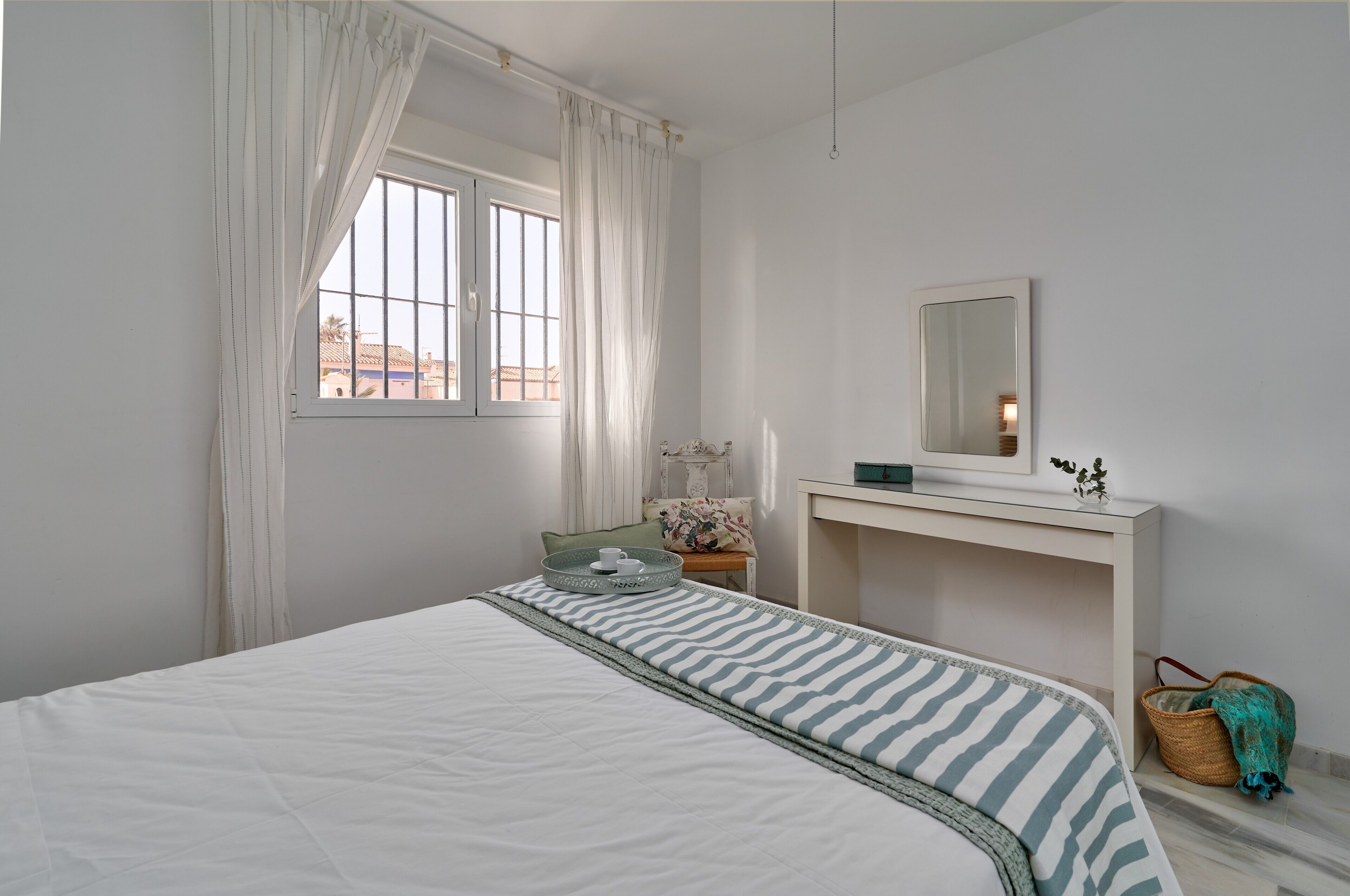 Ein Schlafzimmer mit Bett, Nachttisch, Spiegel, Fenster mit Vorhängen und Blick auf Gebäude.