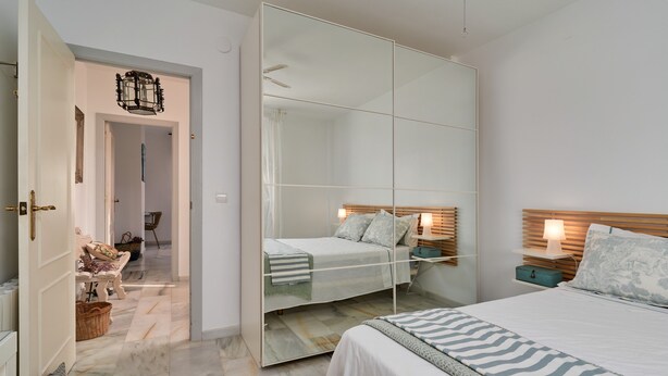 Une chambre avec un grand miroir, un lit recouvert d’une couvre-lit à rayures, une table de chevet avec une lampe et une tête de lit en bois.