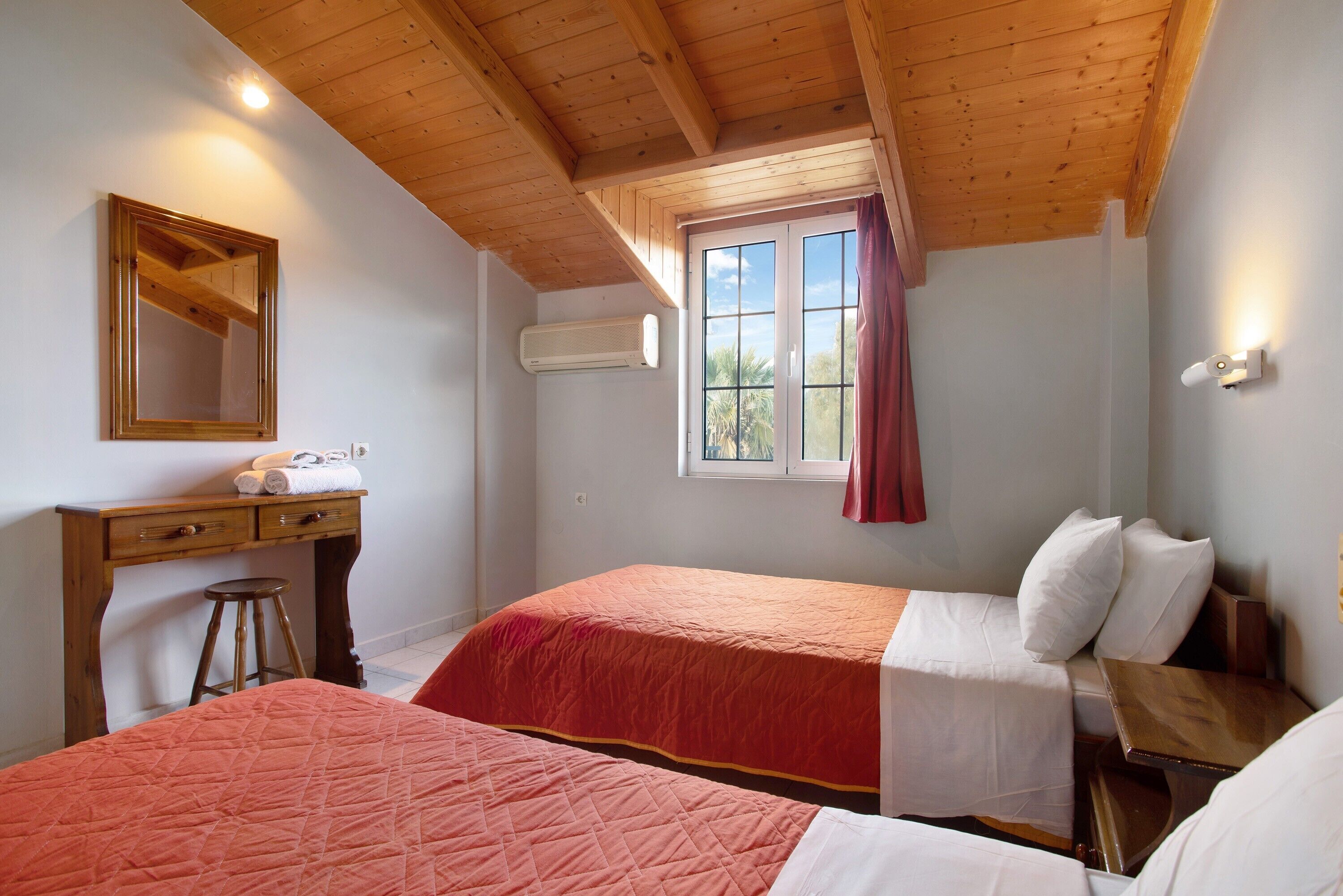 Ein Schlafzimmer mit zwei Betten, Holzboden, Fenster mit Vorhängen, Schreibtisch mit Spiegel und Klimaanlage.