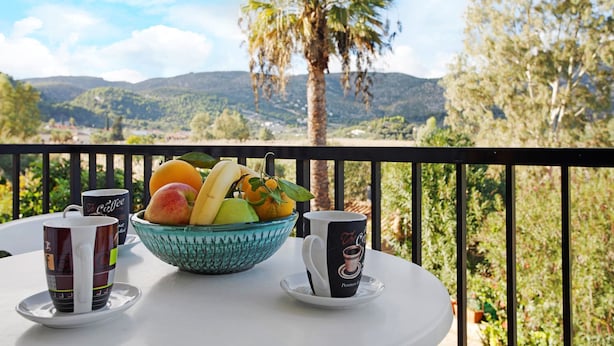 Et bord med en skål frugt og to kaffekopper på en balkon med udsigt over et bjerrelandskab.