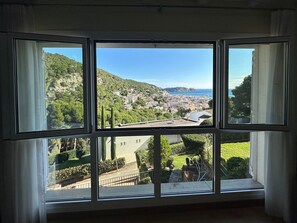 Een kamer met een groot raam, met uitzicht op een kustplaats en heuvels.