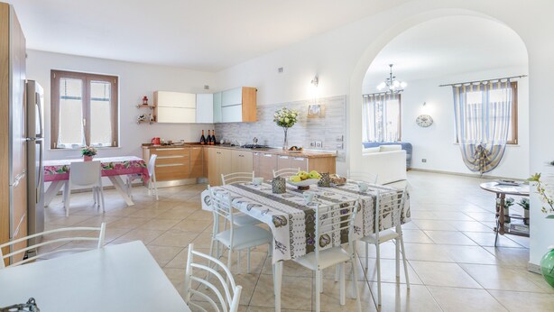 Une cuisine moderne avec des meubles blancs, un coin repas comprenant une table et des chaises, et un espace de vie avec un canapé et des rideaux.