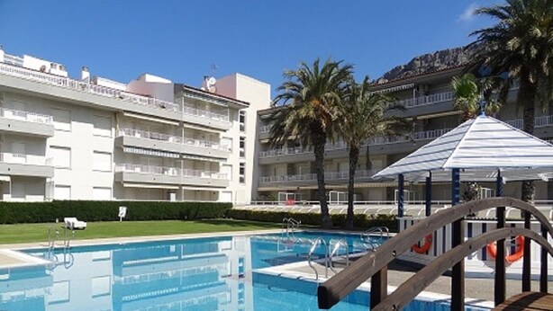 A swimming pool with a wooden deck, surrounded by palm trees and a multi-story building with balconies. - Holiday Apartment "Mar D'or 223" with Shared Pool, Terraces & Wi-Fi (girona)