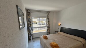 A hotel room with a bed, a window with curtains, a small table, and a wall-mounted light. - Holiday Apartment "Mar D'or 223" with Shared Pool, Terraces & Wi-Fi (girona)