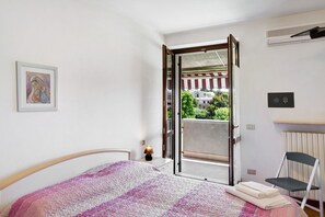 Une chambre à coucher avec un lit, une chaise, un radiateur, un tableau accroché au mur et une porte ouverte donnant sur un balcon.