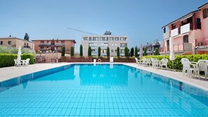 Ein Schwimmbecken mit gefliestem Boden, umgeben von weißen Plastikstühlen und -tischen, mit Gebäuden im Hintergrund.