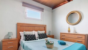 Une chambre à coucher avec une tête de lit en bois, une fenêtre avec des stores, un miroir rond, une table de chevet avec une lampe, une commode, un lit avec une couverture bleue et un panier contenant un ours en peluche.