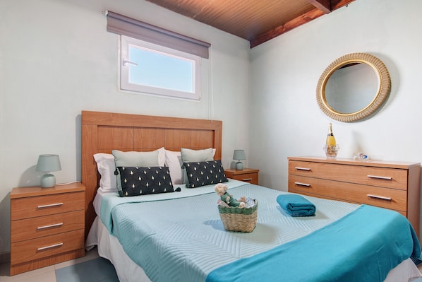 A bedroom with a wooden headboard, a window with blinds, a round mirror, a bedside table with a lamp, a nightstand, a bed with a blue blanket, and a basket with a teddy bear.