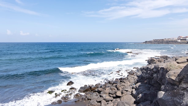 Une côte rocheuse où les vagues s’écrasent sur le rivage, un ciel dégagé et des bâtiments en arrière-plan.