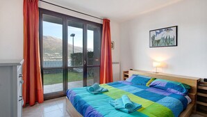 Une chambre avec un grand lit, une porte-fenêtre coulissante donnant sur l’extérieur, une table de chevet avec une lampe et un tableau encadré au mur.