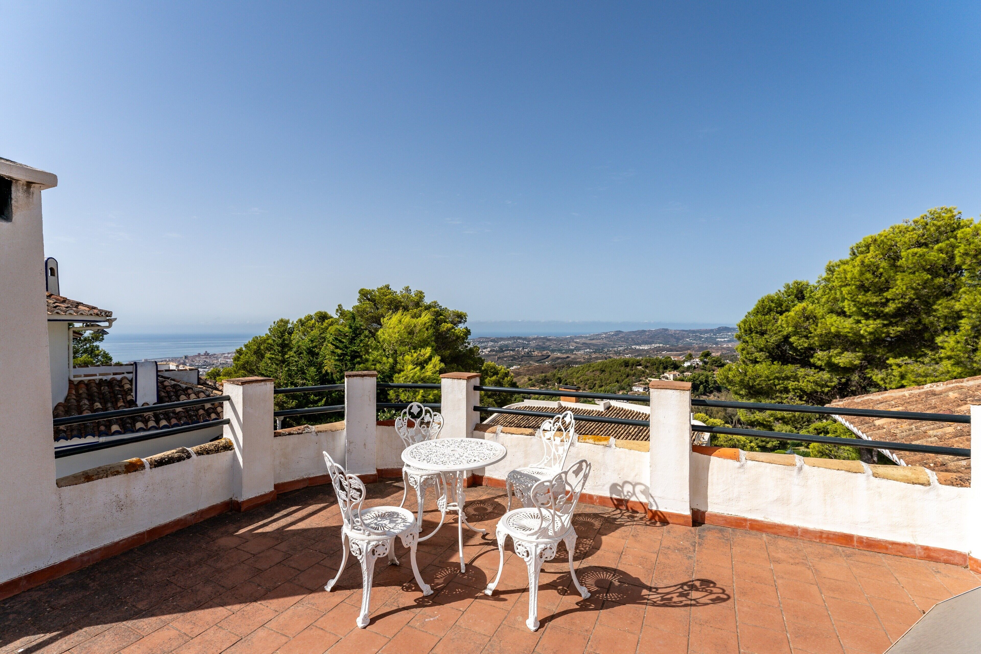 Una terraza con sillas y mesa de metal blanco, con vista a un paisaje pintoresco con árboles y una vista distante del mar.