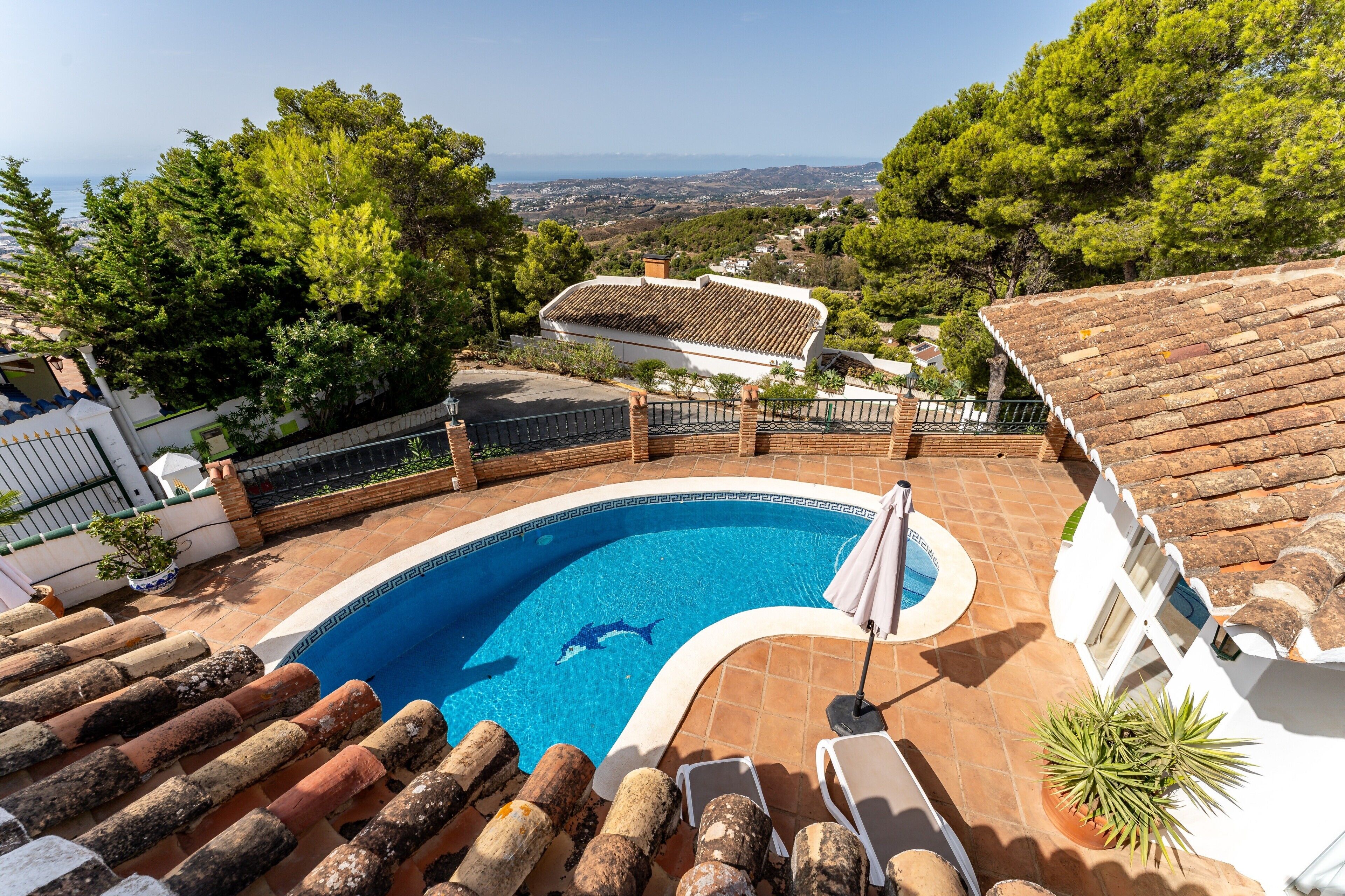 Una piscina con un delfín, rodeada por un edificio blanco con tejado de azulejos, y vistas al paisaje.
