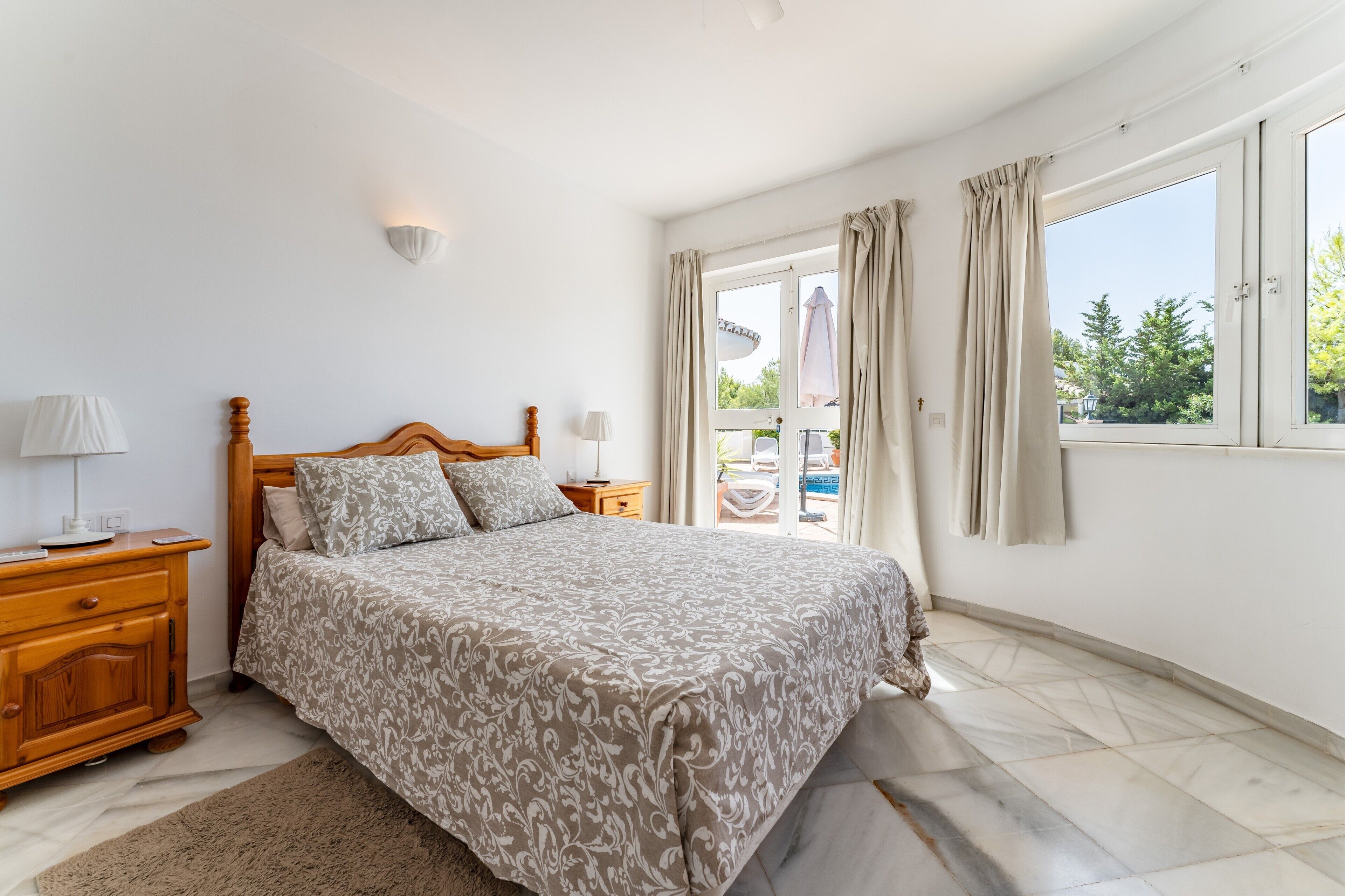 Un dormitorio con cama, mesitas de noche, lámpara, ventana con cortinas y vistas al exterior.