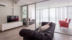 En moderne stue med et stort fladskærms-tv, en mørk sofa og en glasvæg, der fører ud til en balkon med røde stole.