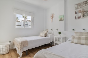 Quarto com duas camas de solteiro, uma mesa pequena, parede branca com um desenho de árvore e janela com vista para edifícios.