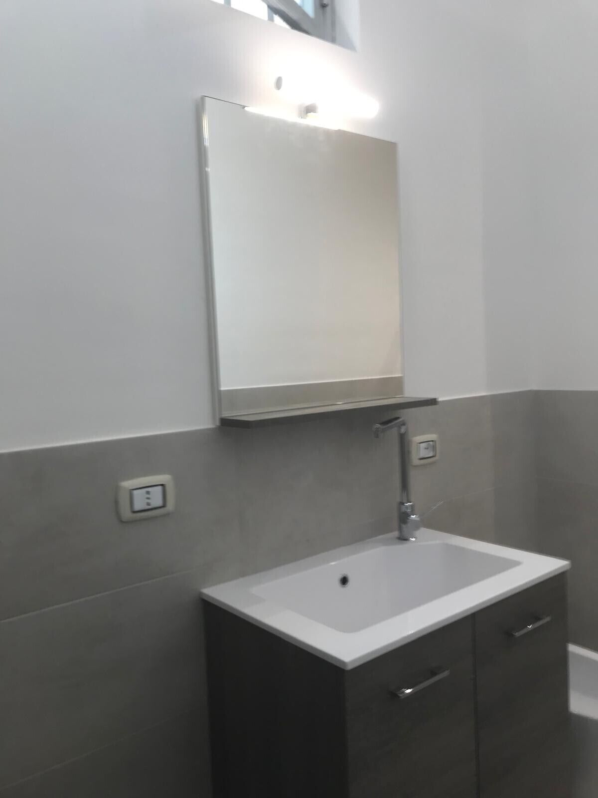 Un baño con lavamanos blanco, muebles oscuros, un espejo y una lámpara sobre el espejo.