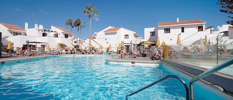 A swimming pool with lounge chairs and umbrellas, surrounded by white buildings with red-tiled roofs.