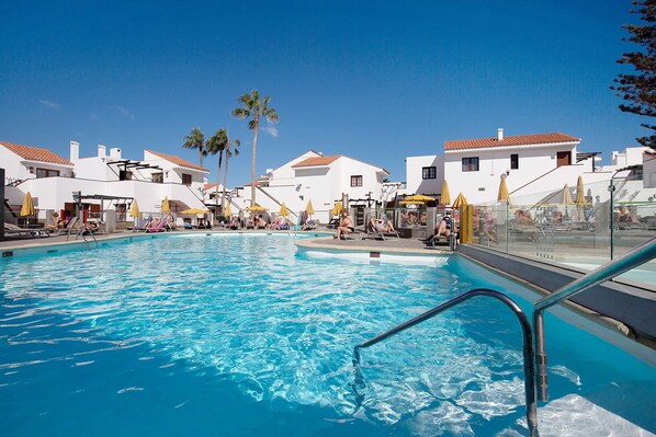 A swimming pool with lounge chairs and umbrellas, surrounded by white buildings with red-tiled roofs.