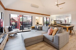 A spacious living area with a large grey sectional sofa, a wooden coffee table, and a TV stand. A dining area with a table and chairs is visible in the background. - CONVENIENTLY LOCATED 250M (APPROX) TO THE EXQUISITE SHORES OF GEOGRAPHE BAY (Dunsborough)