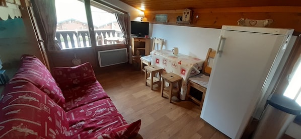 A cozy room with a red sofa, a dining table with chairs, a television, and a refrigerator.