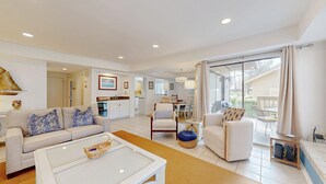 A spacious living area with a sofa, armchairs, and a coffee table. There is a dining area with chairs and a table. The room has a large window with curtains, and a kitchen is visible in the background.