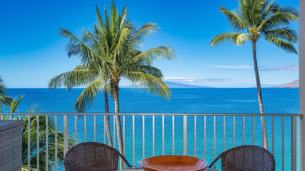 A balcony with two wicker chairs and a table, overlooking a clear blue sea with palm trees. - TOP FLOOR-STUNNING OCEAN VIEWS-WELL EQUIPPED-ALL NEW FURNISHINGS (Kihei)