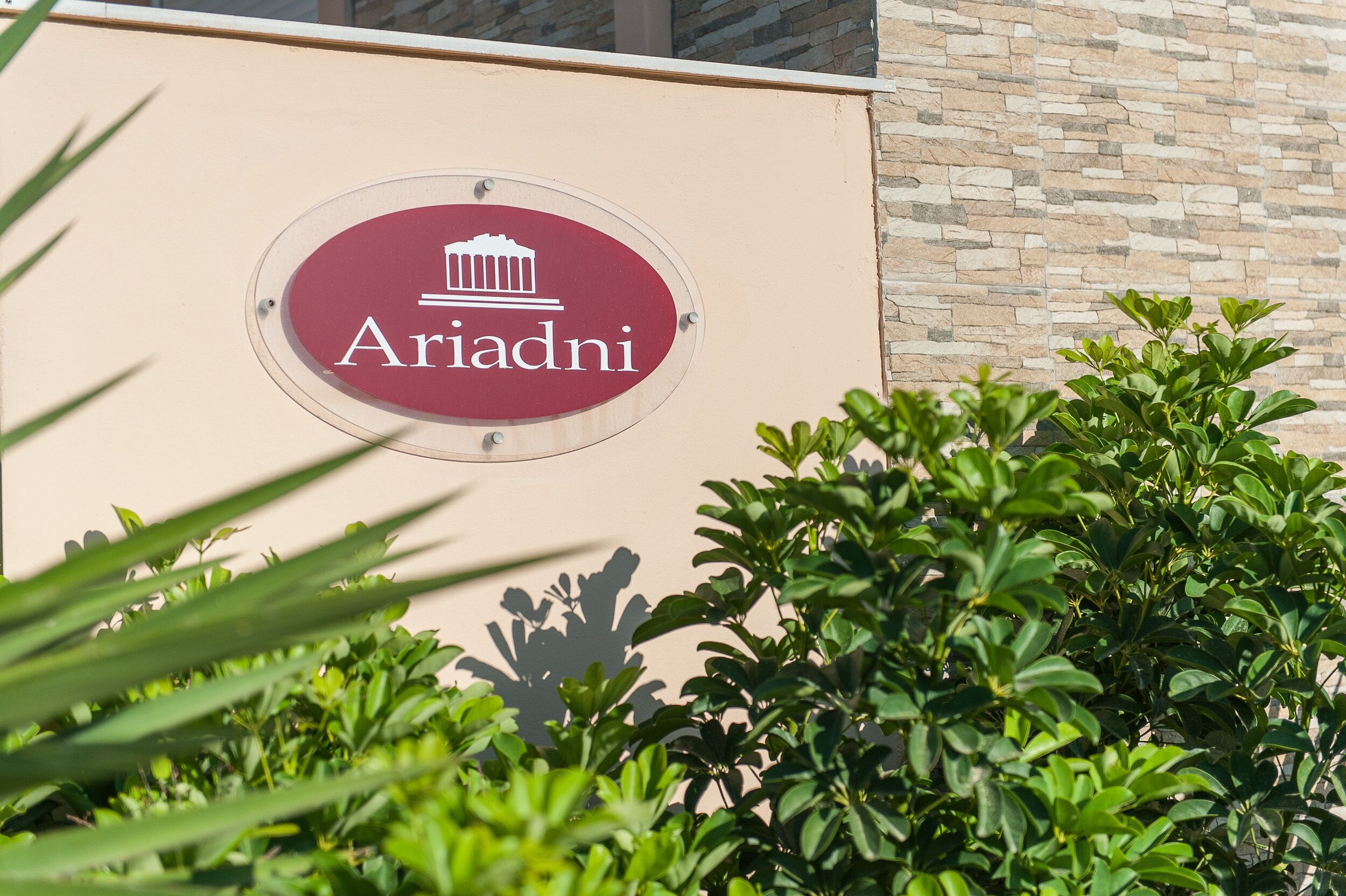 A sign with the name "Ariadni" and a building facade with a stone wall and greenery.