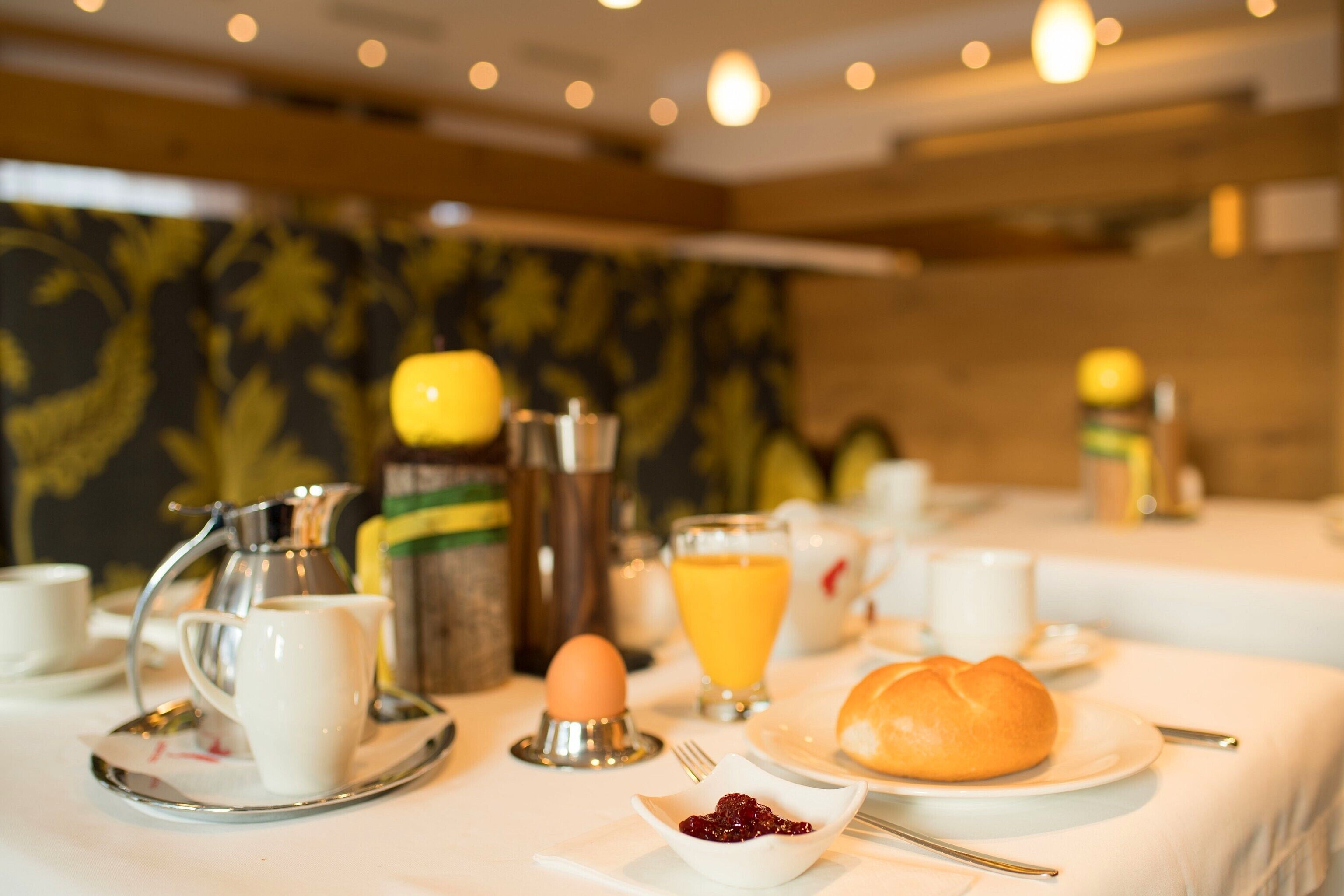 A breakfast setting with a coffee pot, egg, orange juice, croissant, and utensils on a table.