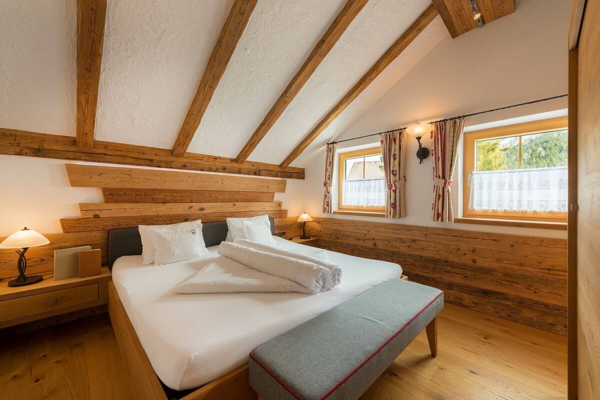 A cozy bedroom with a wooden ceiling, a large bed with white linens, a bedside table with a lamp, and a window with curtains.