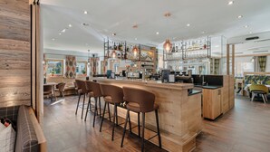 Zona de bar moderna con mostrador de madera, taburetes y una variedad de botellas y vasos en estanterías.