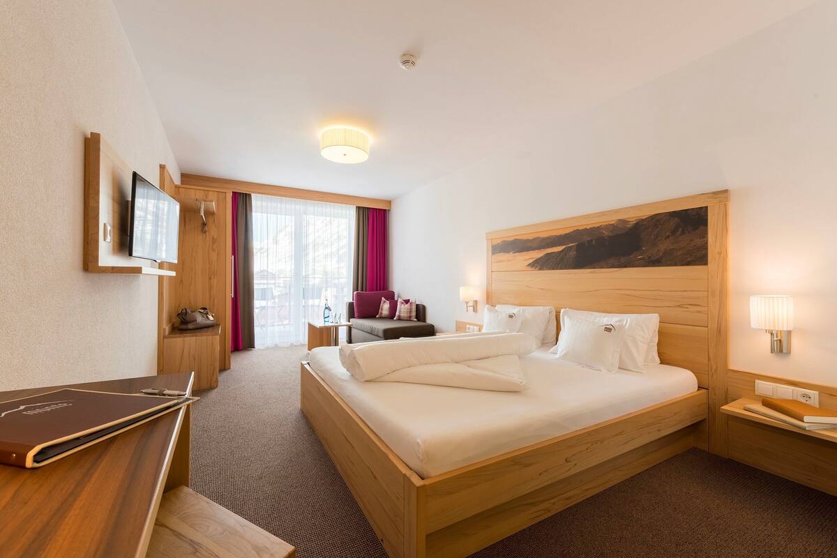 A hotel room with a wooden bed, a desk with a laptop, a television mounted on the wall, and a large painting above the bed.
