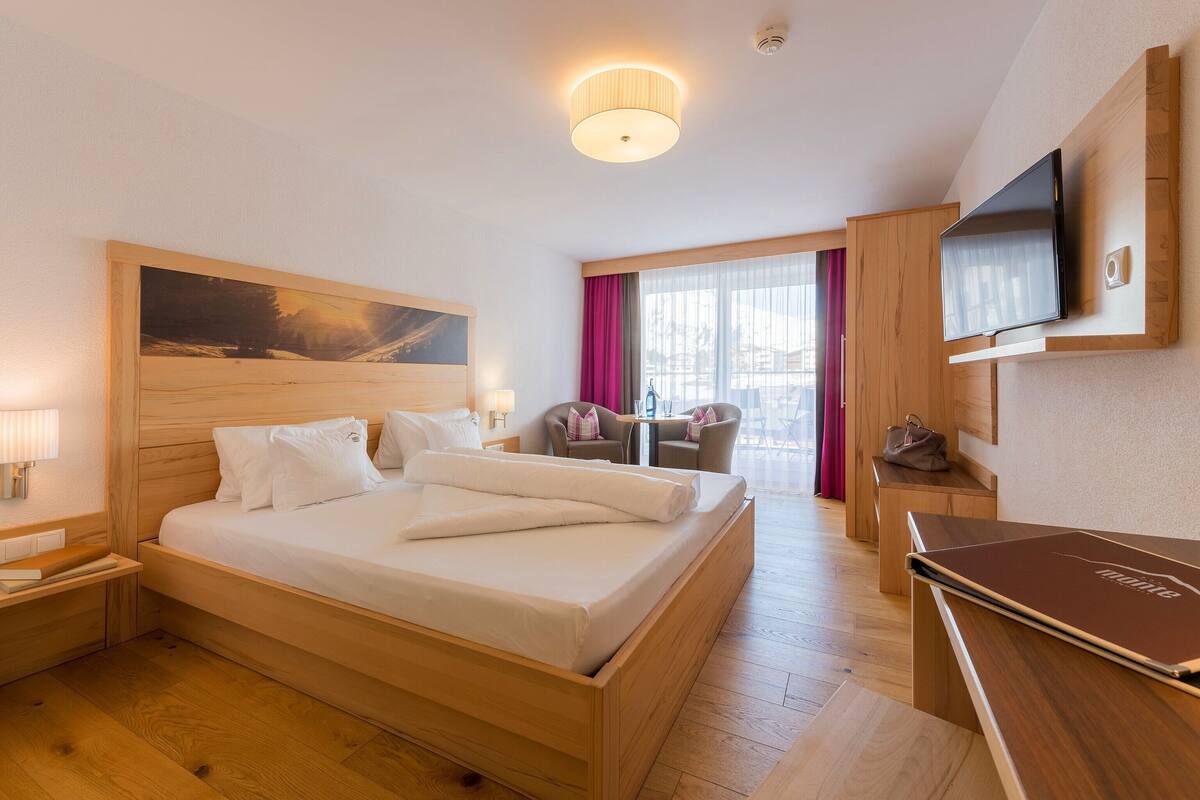 A hotel room with a large bed, wooden headboard, a flat-screen TV mounted on the wall, a desk with a chair, and a window with curtains.