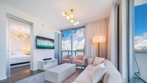Una habitación de hotel moderna con cama, sofá, mesa de centro, televisor y un amplio ventanal con vistas.