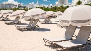 Una playa con filas de tumbonas y sombrillas, un cielo despejado y edificios al fondo.