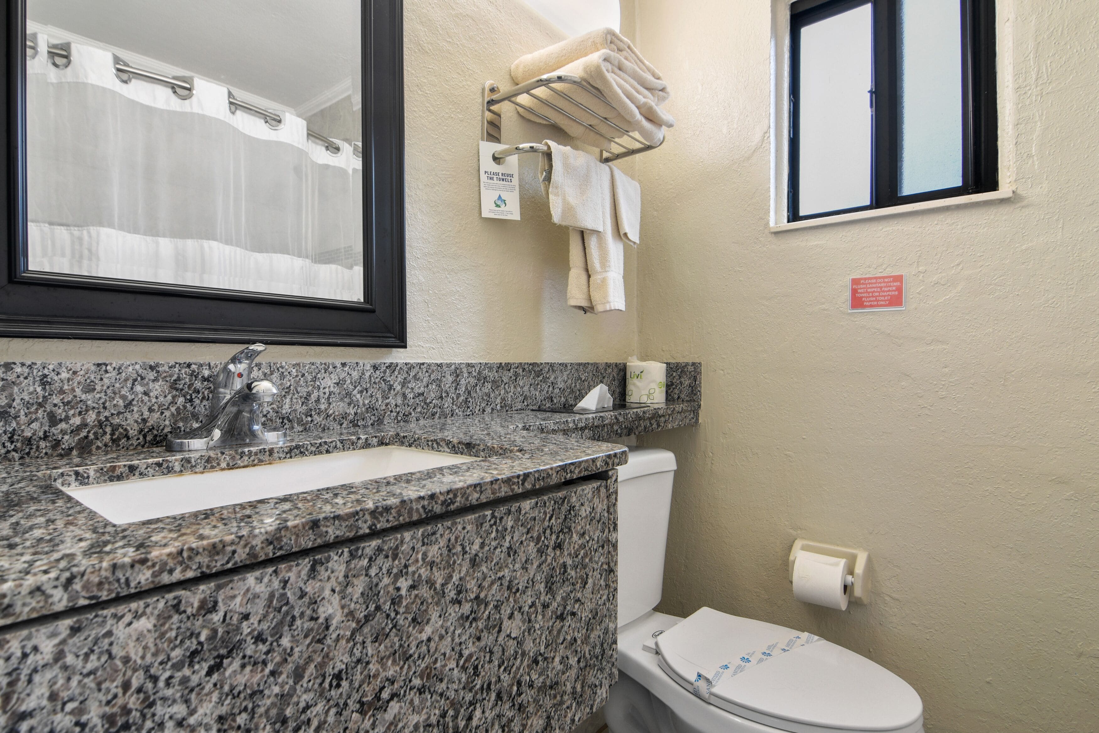A bathroom with a granite countertop sink, a mirror, a towel rack with folded towels, a toilet, and a window with a view of the outside.