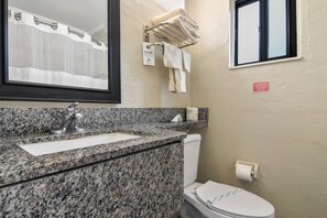 A bathroom with a granite countertop sink, a mirror, a towel rack with folded towels, a toilet, and a window with a view of the outside. - Anthony's on the Beach (Cocoa Beach)
