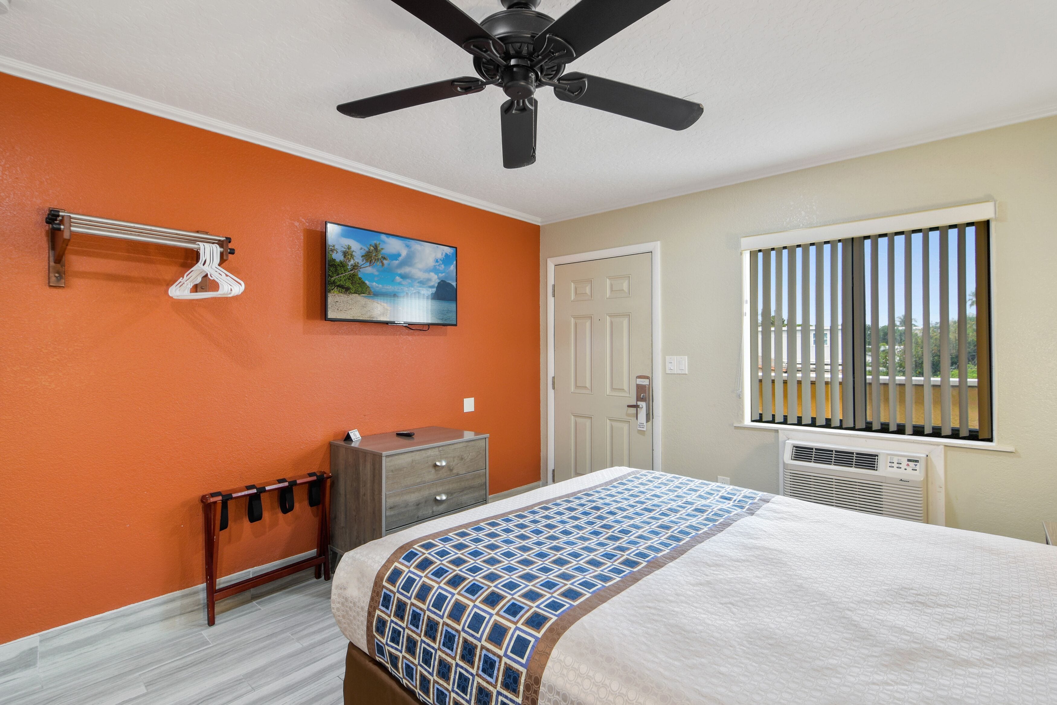 Un dormitorio con pared naranja, cama con colcha estampada, ventilador de techo, cómoda, perchero y ventana con persianas.