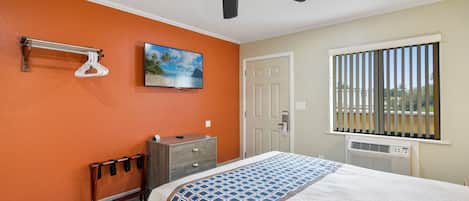 Une chambre avec un mur orange, un lit recouvert d’une couvre-lit à motifs, un ventilateur de plafond, une commode, un porte-manteau et une fenêtre avec des stores.