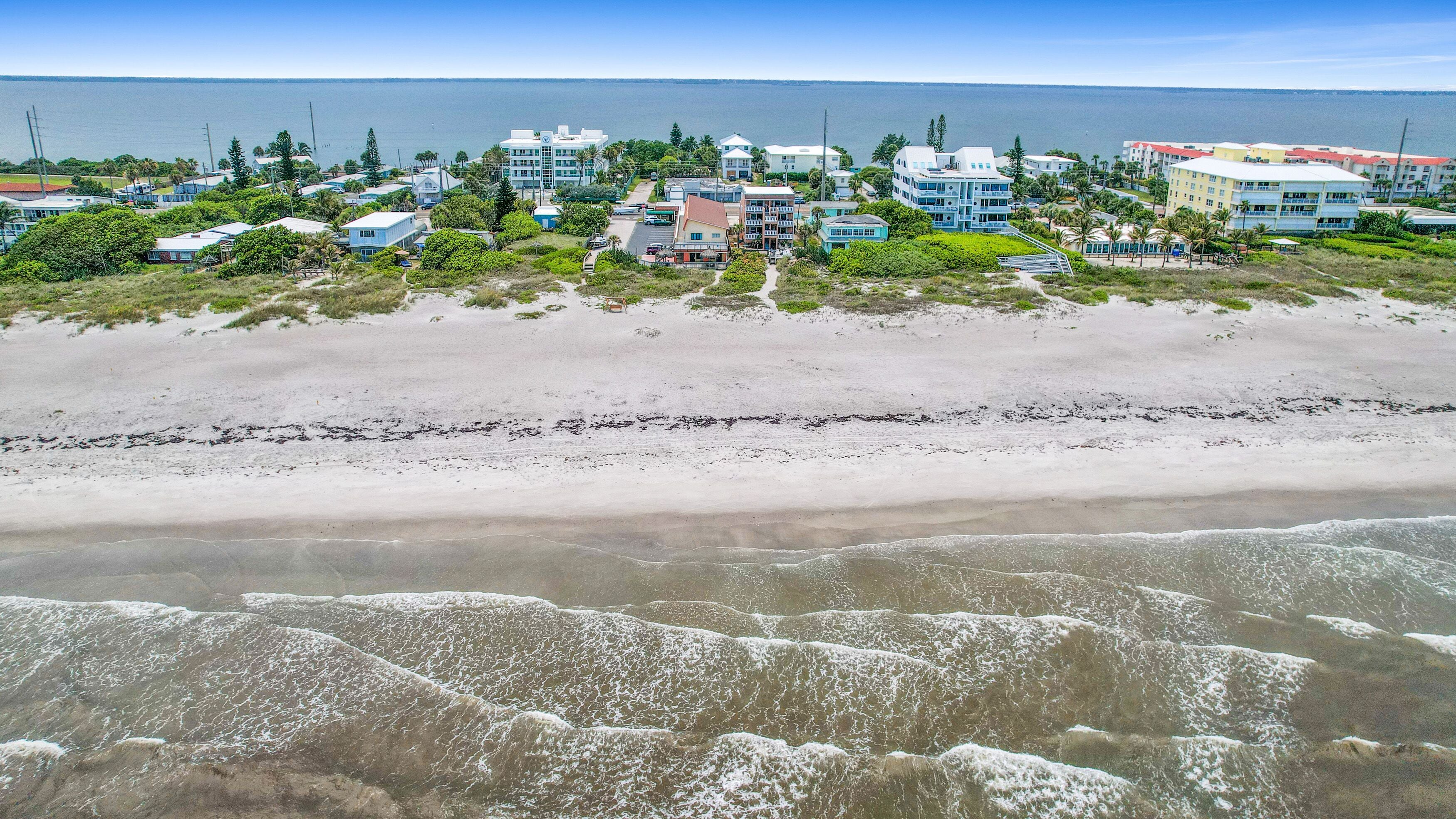 A coastal area with beachfront properties, including multi-story buildings and single-family homes, with a sandy beach and ocean in the background.