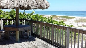 Una terraza de madera con un toldo de paja, rodeada por barandillas de madera, con vistas a una playa con dunas y el océano.