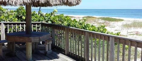 Una terraza de madera con un toldo de paja, rodeada por barandillas de madera, con vistas a una playa con dunas y el océano.