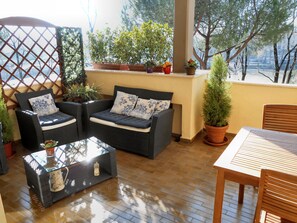 A covered patio with two dark-colored sofas, a glass coffee table, and a wooden table. There are potted plants and a view of trees and a body of water in the background. - Riverfront Terrace, 3 Bedrooms, 3 Bathrooms, Garage (Firenze)