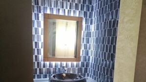 Une salle de bain avec un mur recouvert de carreaux de marbre, un meuble-lavabo en bois et un lavabo rond posé sur le plan de toilette.
