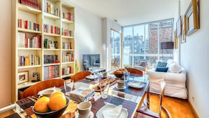 A modern living room with a dining area, large windows, a bookshelf, and a white sofa.
