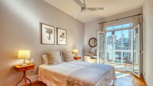 A bedroom with a bed, bedside tables, a ceiling fan, a window with a view of buildings, and framed pictures on the wall.